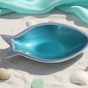May include: A fish-shaped decorative bowl with a vibrant teal interior and a silver-colored exterior. The bowl is set on a bed of white sand, with seashells, small stones, and a piece of coral nearby. A light blue fabric drapes in the background.