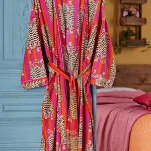 May include: A vibrant pink and orange striped robe with a tiger print. The robe has a matching belt and white trim. It is hanging on a wooden hanger against a blue door. The robe is approximately 55 inches long.