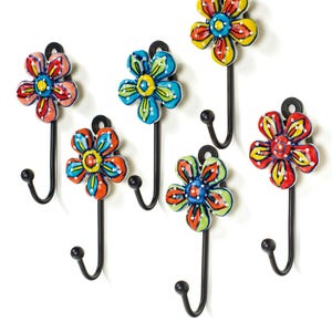 May include: A set of six wall hooks, each featuring a hand-painted ceramic flower head in vibrant colours including red, yellow, blue, and green. Each flower is attached to a black metal hook with a rounded tip, designed for mounting on a wall.
