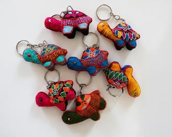May include: A collection of colorful, embroidered turtle keychains. Each keychain features a unique design with vibrant hues of red, blue, green, and yellow. The keychains are made of fabric and have metal key rings.