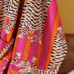 May include: A vibrant, striped robe featuring a tiger print. The robe has a pink, orange, and white color scheme. The tiger print is black and tan. The robe has a white trim and a matching belt.