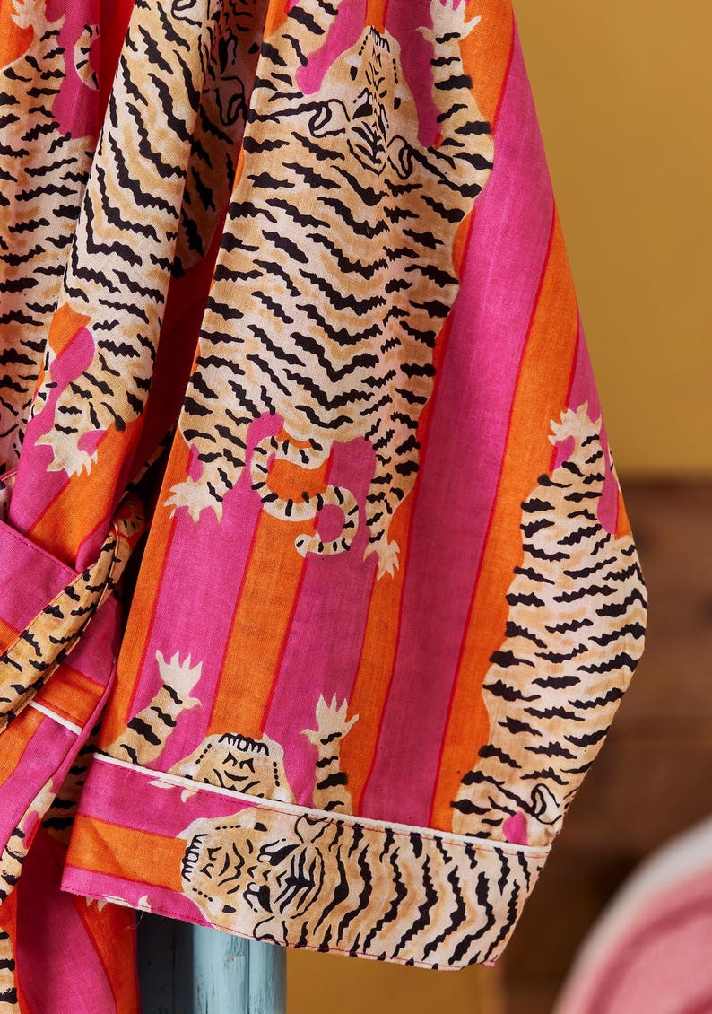 May include: A vibrant, striped robe featuring a tiger print. The robe has a pink, orange, and white color scheme. The tiger print is black and tan. The robe has a white trim and a matching belt.
