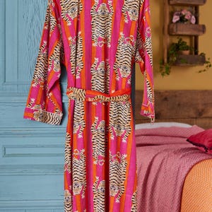 May include: A vibrant pink and orange striped robe with a tiger print design. The robe is hanging on a wooden hanger. The robe has a matching belt and white trim.