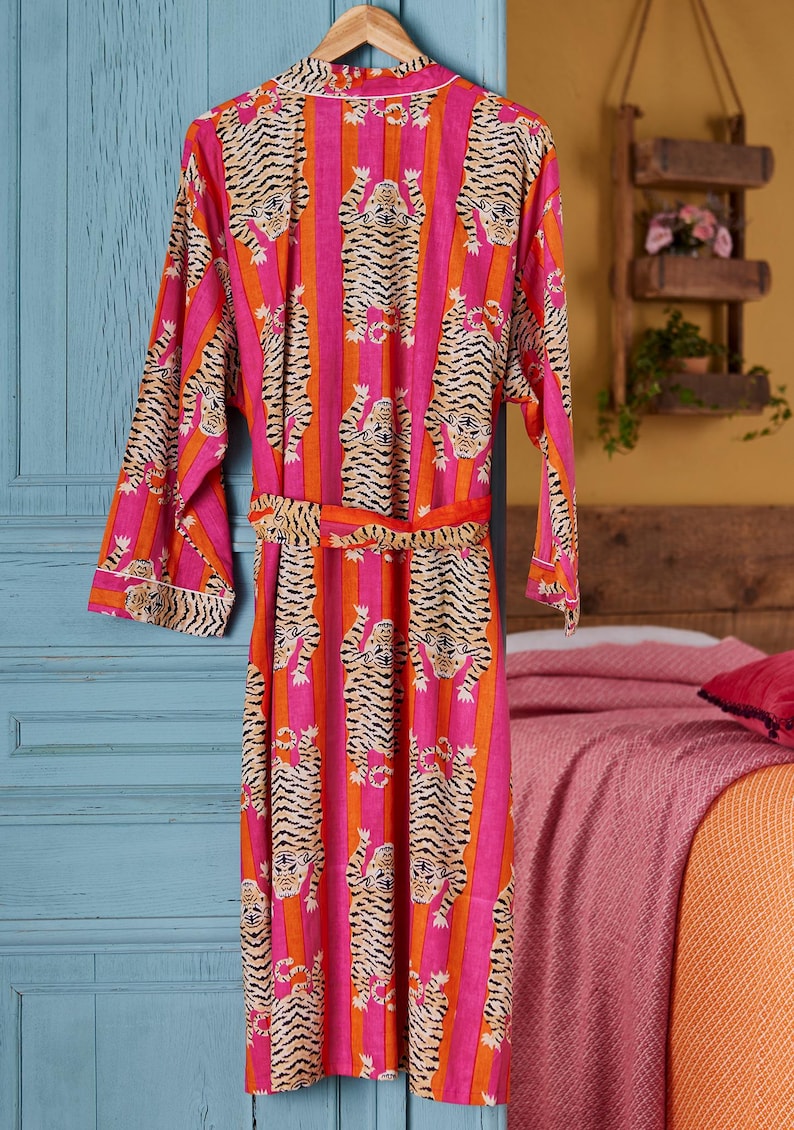 May include: A vibrant pink and orange striped robe with a tiger print design. The robe is hanging on a wooden hanger. The robe has a matching belt and white trim.