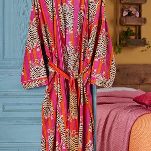 May include: A vibrant pink and orange striped robe with a tiger print design. The robe has a matching belt and white trim. It is hanging on a wooden hanger against a blue door. The robe is approximately 55 inches long.
