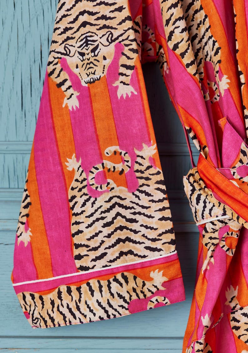 May include: A vibrant, patterned robe featuring a tiger print. The robe has a pink, orange, and tan color scheme with vertical stripes and tiger illustrations. The sleeves and trim are detailed with white borders. The robe is tied at the waist.
