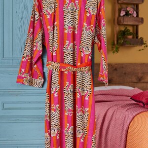 May include: A vibrant pink and orange striped robe with a tiger print design. The robe is hanging on a wooden hanger. The robe has a matching belt and white trim.