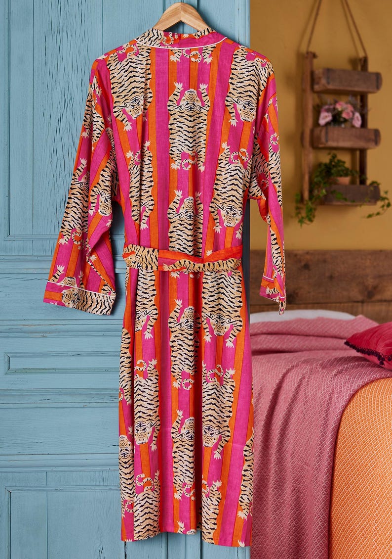 May include: A vibrant pink and orange striped robe with a tiger print design. The robe is hanging on a wooden hanger. The robe has a matching belt and white trim.