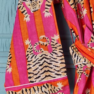 May include: A vibrant, patterned robe featuring a tiger print. The robe has a pink, orange, and tan color scheme with vertical stripes and tiger illustrations. The sleeves and trim are detailed with white borders. The robe is tied at the waist.