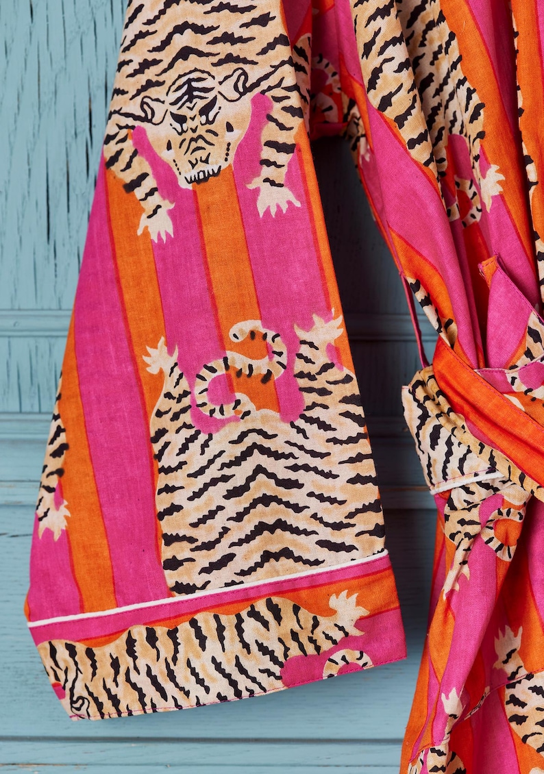 May include: A vibrant, patterned robe featuring a tiger print. The robe has a pink, orange, and tan color scheme with vertical stripes and tiger illustrations. The sleeves and trim are detailed with white borders. The robe is tied at the waist.