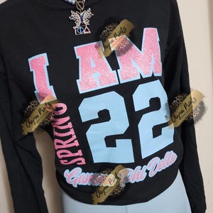 Peut inclure: T-shirt à manches longues noire avec un motif "I AM" en paillettes roses et "22" en bleu. Le t-shirt comporte également le texte "Spring 2022 Gamma Phi Delta" en paillettes roses. Le t-shirt est porté par un mannequin portant un collier de perles bleu clair avec un pendentif en forme de papillon.