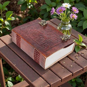 Puede incluir: Un diario rectangular encuadernado en cuero de color marrón oscuro, con un diseño de árbol en relieve en la cubierta y dos cierres metálicos de estilo antiguo. El diario tiene páginas blancas gruesas y costuras decorativas en el lomo.
