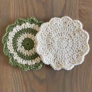 Op de afbeelding: Twee handgemaakte gehaakte onderzetters. Eén onderzetter heeft een groen-wit spiraalontwerp met een geschulpte rand. De andere onderzetter is effen wit met een geschulpte rand. Beide onderzetters zijn cirkelvormig en liggen op een houten oppervlak.