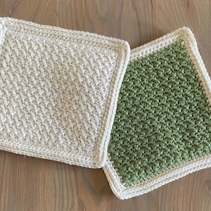 Op de afbeelding: Twee vierkante gehaakte pannenlappen. Eén is gebroken wit, de andere is groen. Beide hebben een gestructureerd steekpatroon en een witte rand. De pannenlappen zijn handgemaakt en geschikt voor gebruik in de keuken.