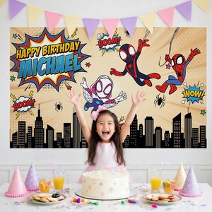 May include: A birthday party scene with a backdrop featuring Spider-Man characters and the text "Happy Birthday Michael." The backdrop is tan with comic book style graphics. A young girl is in front of the backdrop, smiling with her arms raised. A cake and party supplies are on the table.