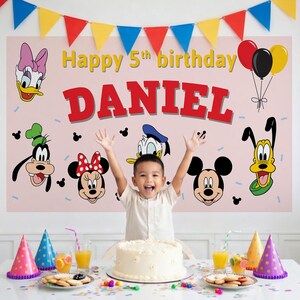 May include: A child celebrates their 5th birthday in front of a pink banner with the name "DANIEL" in red letters. The banner features cartoon characters and the text "Happy 5th birthday". A cake, party hats, and balloons are also visible.