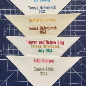 May include: Four white triangle fabric patches with embroidered text. The top patch reads "The Mountains are Calling" with the name "Teresa Hafenbrack" and the year "2024" below. The second patch reads "Scattered Leaves" with the name "Teresa Hafenbrack" and the year "2024" below. The third patch reads "Heaven and Nature Sing" with the name "Teresa Hafenbrack" and the date "July 2024" below. The bottom patch reads "Tulip Season" with the name "Connie Little" and the year "2024" below.