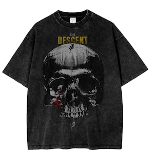 May include: A charcoal grey acid-wash t-shirt featuring a large graphic of a human skull. Above the skull is the text THE DESCENT in yellow. The skull design includes small figures of climbers, with a red accent near the eye socket.
