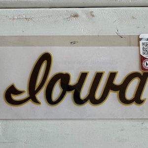 May include: A gold vinyl decal with the word "Iowa" in a cursive font. The decal is packaged in a clear plastic bag with a small sticker on the bottom left corner that says "by Designs by Dawn".