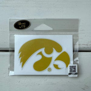 May include: A gold-colored sticker featuring the Iowa Hawkeyes logo, packaged in a clear plastic sleeve. The sticker has a white background and a registered trademark symbol. The packaging includes a black and gold oval with the text "Designs by CJ".