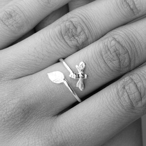 Op de afbeelding: Een zilveren ring met een blad- en bijendesign. De ring wordt gedragen aan de middelvinger van een hand.