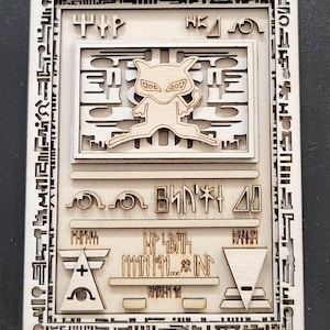 Könnte beinhalten: Eine Holzkarte mit Laserzuschnitt, die ein stilisiertes katzenartiges Wesen in der Mitte zeigt. Die Karte hat einen komplizierten Rand und verschiedene Symbole und Texte. Das Design ist in hellbraunen und beigen Farbtönen gehalten, mit einem geschichteten Effekt.