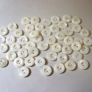 May include: A collection of round, off-white buttons with two holes each. The buttons have a slightly iridescent sheen, suggesting a mother-of-pearl or similar material. They are scattered across a light-colored surface, providing a neutral backdrop.