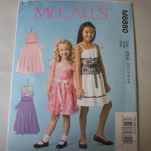 May include: A McCall's sewing pattern, M6880, featuring illustrations of girls' dresses. The pattern includes designs for various styles, including a pink dress with a ribbon, a white dress with black lace, and a purple dress. The size range is CHJ (7-8-10-12-14).