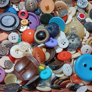May include: A large collection of assorted sewing buttons in various shapes, sizes, and colors including blue, orange, brown, white, black, and purple. The buttons feature different textures, hole counts, and patterns, creating a dense pile.