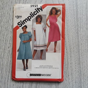 May include: Simplicity sewing pattern number 5921 for a dress. The pattern shows three different styles of dresses: a blue dress with puffed sleeves, a white dress with puffed sleeves and a trim, and a pink sleeveless dress with a tie at the waist. The pattern is labeled "Overnight Success".