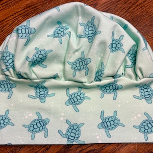 May include: A light mint green scrub cap featuring a repeating pattern of dark teal sea turtles. The hat has a gathered, rounded crown and a wide, flat band that encircles the head, showcasing a consistent print of swimming turtles.