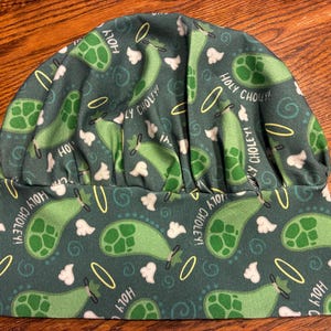 May include: A dark green scrub cap featuring a repeating pattern of light green cartoon pickles with yellow halos and white clouds. The fabric includes the text Holy Choley! printed in white throughout the design. The cap has a gathered top.