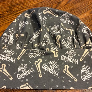 May include: A dark gray scrub cap featuring a repeating pattern of white text that reads "WE NAILED YOUR GRANDMA" interspersed with illustrations of nails and small cross symbols. The cap has a wide, flat band and a gathered, rounded top section.