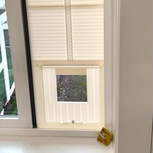 Op de afbeelding: Een wit raam met een cr&egrave;mekleurige geplooide rolgordijn en een transparante kattenluik ge&iuml;nstalleerd. Een kleine gele doos met tekst staat op de vensterbank. De kattenluik is rechthoekig met een vierkante opening.