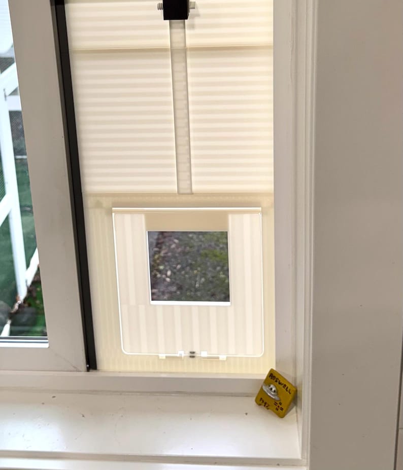 Op de afbeelding: Een wit raam met een cr&egrave;mekleurige geplooide rolgordijn en een transparante kattenluik ge&iuml;nstalleerd. Een kleine gele doos met tekst staat op de vensterbank. De kattenluik is rechthoekig met een vierkante opening.