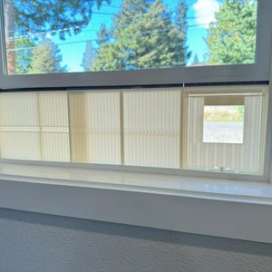 K&ouml;nnte beinhalten: Ein wei&szlig; gerahmtes Fenster mit Blick auf B&auml;ume und Himmel. Das Fenster hat beige vertikale Jalousien, die das Glas teilweise bedecken. Ein kleiner Teil der Jalousien ist ge&ouml;ffnet und gibt den Blick auf eine Stra&szlig;e und Gr&uuml;n frei.