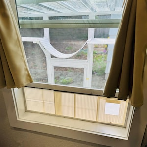 K&ouml;nnte beinhalten: Ein wei&szlig; gerahmter Katzenk&auml;fig mit einer H&auml;ngematte im Inneren, sichtbar durch ein Fenster. Beige Vorh&auml;nge umrahmen das Fenster. Das Gehege hat ein Maschendach und einen wei&szlig;en Rahmen. Das Fenster hat einen wei&szlig;en Rahmen und eine beige Jalousie.