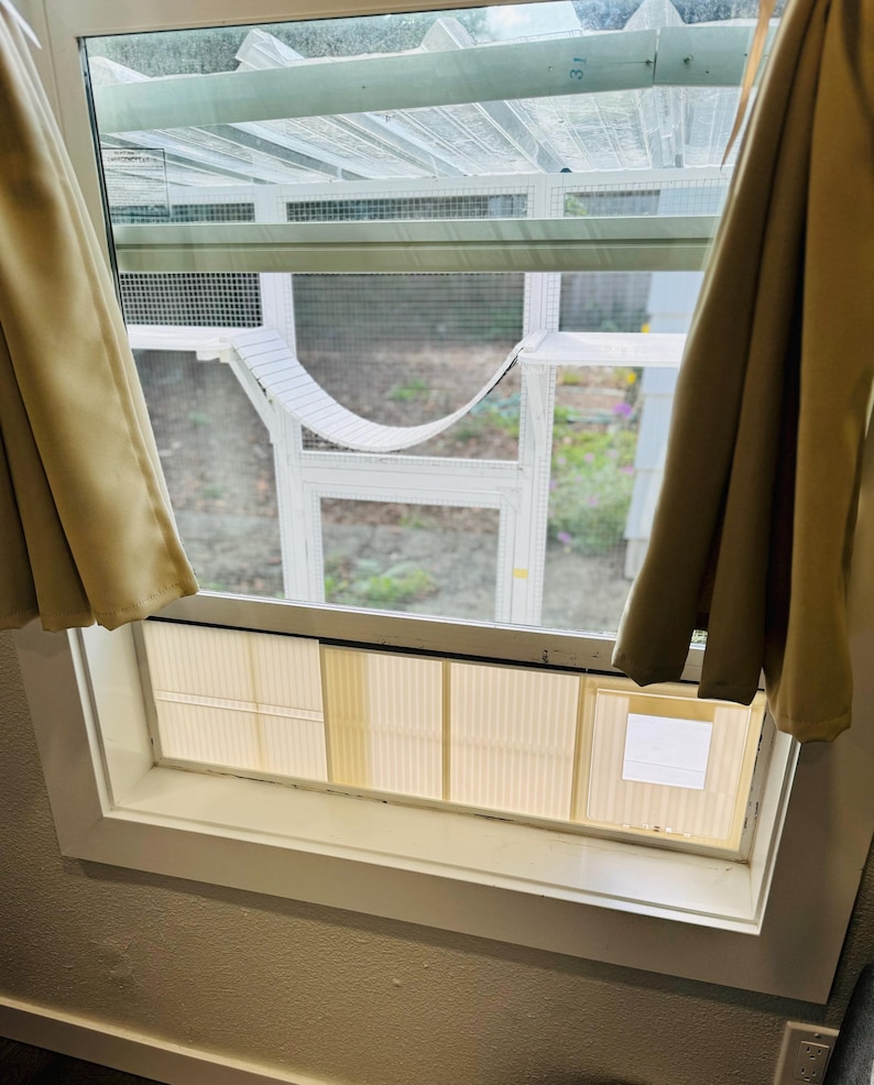K&ouml;nnte beinhalten: Ein wei&szlig; gerahmter Katzenk&auml;fig mit einer H&auml;ngematte im Inneren, sichtbar durch ein Fenster. Beige Vorh&auml;nge umrahmen das Fenster. Das Gehege hat ein Maschendach und einen wei&szlig;en Rahmen. Das Fenster hat einen wei&szlig;en Rahmen und eine beige Jalousie.