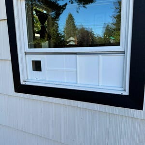 K&ouml;nnte beinhalten: Ein wei&szlig;es Fenster mit schwarzem Rahmen ist in wei&szlig;e Verkleidung eingelassen. Das Fenster spiegelt einen blauen Himmel und B&auml;ume wider. Das Fenster hat eine kleine quadratische &Ouml;ffnung und ein wei&szlig;es Paneeldesign darunter.