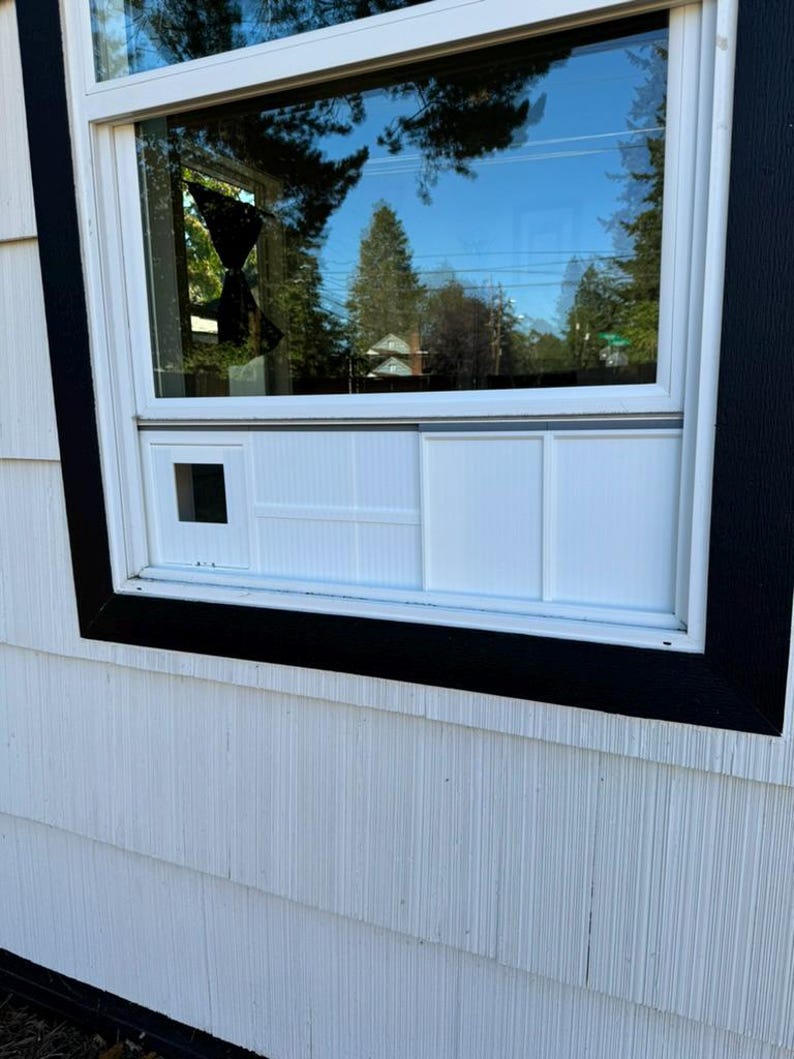 K&ouml;nnte beinhalten: Ein wei&szlig;es Fenster mit schwarzem Rahmen ist in wei&szlig;e Verkleidung eingelassen. Das Fenster spiegelt einen blauen Himmel und B&auml;ume wider. Das Fenster hat eine kleine quadratische &Ouml;ffnung und ein wei&szlig;es Paneeldesign darunter.