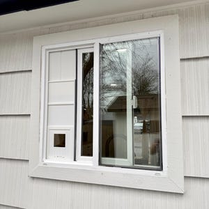 Op de afbeelding: Een witte schuifraam met een ingebouwde kattenluik. Het raam is geplaatst in een wit frame en is ge&iuml;nstalleerd in een wit gevelbekleed gebouw. Het glas weerspiegelt de buitenomgeving, inclusief bomen en een huis.