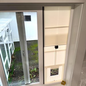 Op de afbeelding: Een raam met een wit frame en een gedeeltelijk geopende glazen schuifdeur. Het raam heeft cr&egrave;mekleurige jaloezie&euml;n en een kleine vierkante opening. Een geel object ligt op de vensterbank.