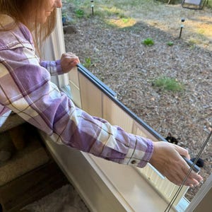 K&ouml;nnte beinhalten: Eine Person in einem lila-wei&szlig; karierten Hemd h&auml;lt ein Fenstergitter. Der Fensterrahmen ist wei&szlig; und der Bildschirm hat einen dunkelgrauen Rahmen. Der Hintergrund zeigt einen Garten mit Liegest&uuml;hlen und einer schwarzen Katze.