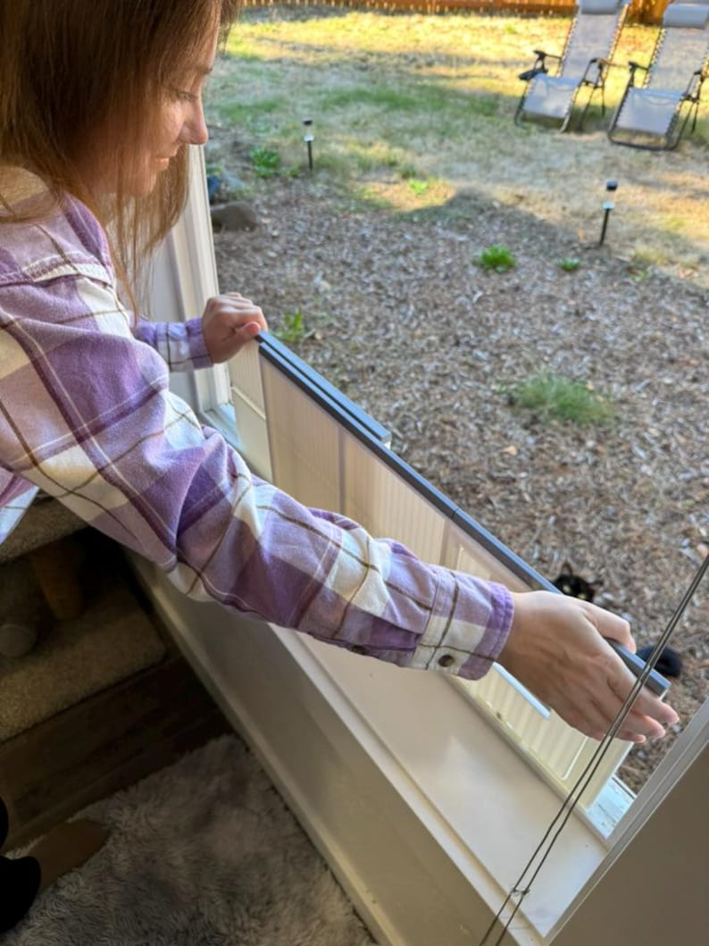 K&ouml;nnte beinhalten: Eine Person in einem lila-wei&szlig; karierten Hemd h&auml;lt ein Fenstergitter. Der Fensterrahmen ist wei&szlig; und der Bildschirm hat einen dunkelgrauen Rahmen. Der Hintergrund zeigt einen Garten mit Liegest&uuml;hlen und einer schwarzen Katze.