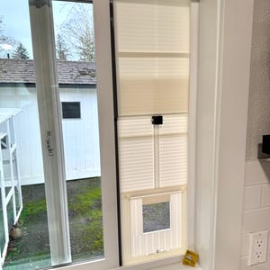 May include: A window with a cream-colored pleated shade and a pet door installed. The window is framed in white and has a view of a backyard with a white shed. A small yellow sticker is on the window frame.