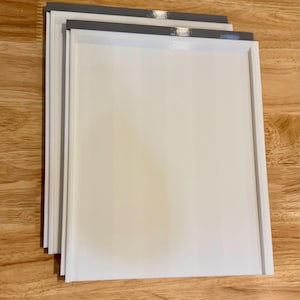 May include: A stack of white rectangular trays with gray trim. The trays are stacked on top of each other, with the top tray slightly offset. The trays are on a wooden surface.