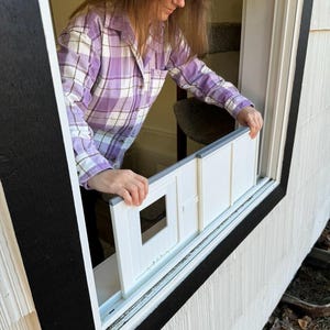 K&ouml;nnte beinhalten: Ein wei&szlig;er Einsatz f&uuml;r eine Katzent&uuml;r wird in ein Fenster eingebaut. Der Einsatz hat eine quadratische &Ouml;ffnung. Eine Person in einem lila karierten Hemd installiert ihn. Der Fensterrahmen ist schwarz, die Hausverkleidung wei&szlig;.