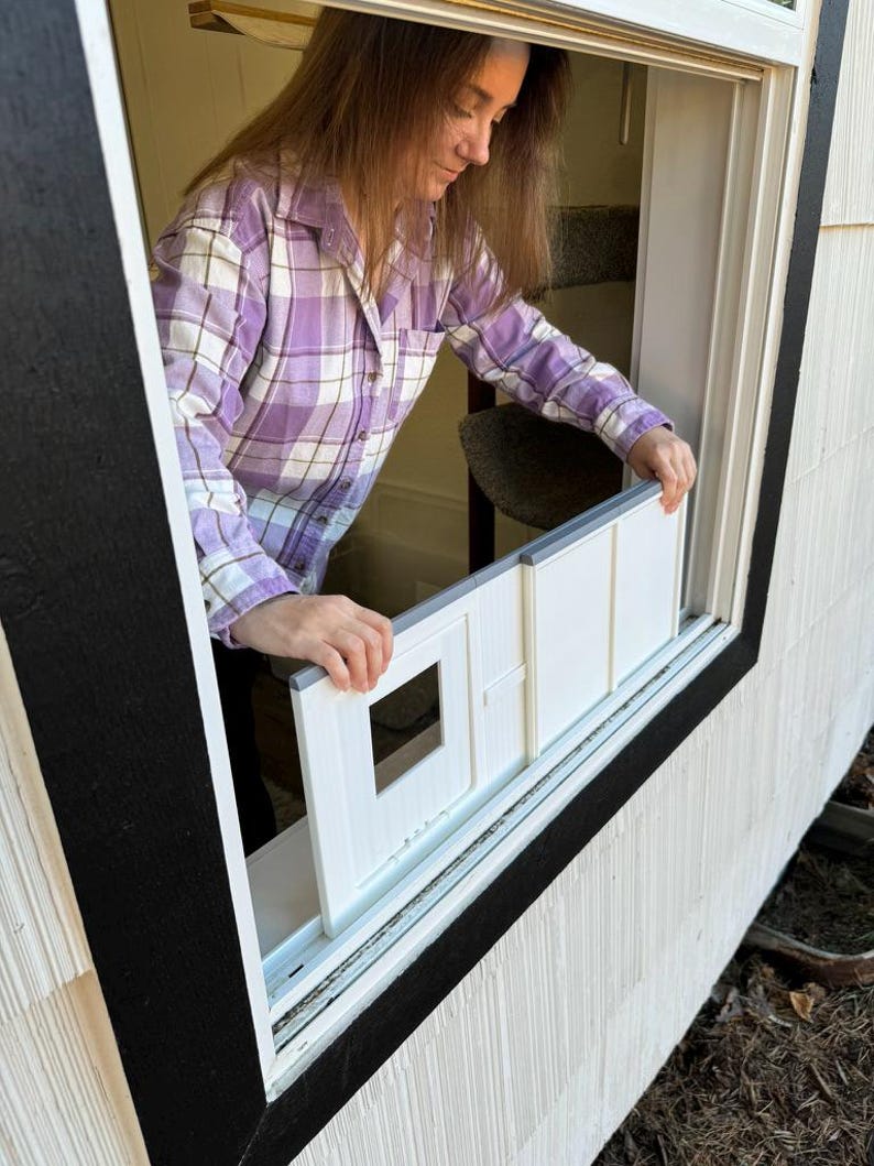 K&ouml;nnte beinhalten: Ein wei&szlig;er Einsatz f&uuml;r eine Katzent&uuml;r wird in ein Fenster eingebaut. Der Einsatz hat eine quadratische &Ouml;ffnung. Eine Person in einem lila karierten Hemd installiert ihn. Der Fensterrahmen ist schwarz, die Hausverkleidung wei&szlig;.