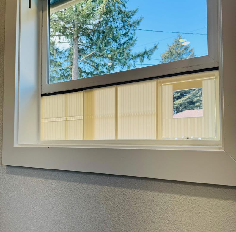 K&ouml;nnte beinhalten: Ein wei&szlig; gerahmtes Fenster mit Blick auf einen Baum und einen blauen Himmel. Das Fenster hat eine hellfarbige, vertikal gestreifte Jalousie. Ein kleines Fenster ist ge&ouml;ffnet und gibt einen Blick nach drau&szlig;en frei.