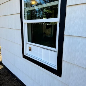 K&ouml;nnte beinhalten: Au&szlig;enansicht eines Hauses mit wei&szlig;er Verkleidung und einem schwarz umrandeten Fenster. Das Fenster spiegelt B&auml;ume und Himmel wider. Das Fenster ist wei&szlig; mit einem schwarzen Rahmen. Die Verkleidung ist strukturiert.